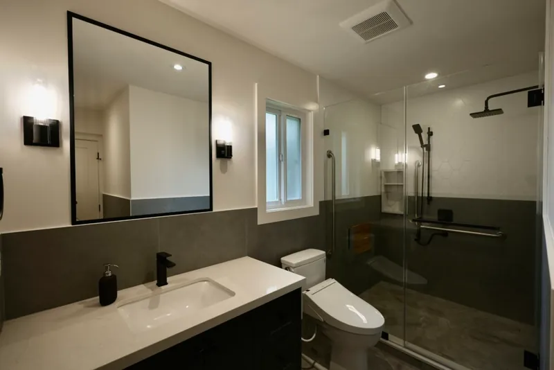 A renovated bathroom with a half-height grey tile wainscot, a glass-screened shower, grab bars and a fold-down seat.