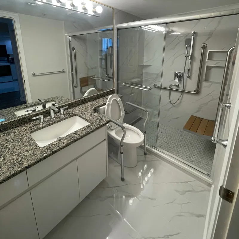 After — A renovated bathroom in marble-look tile with a curbless walk-in shower, grab bars and a fold-down teak bench.